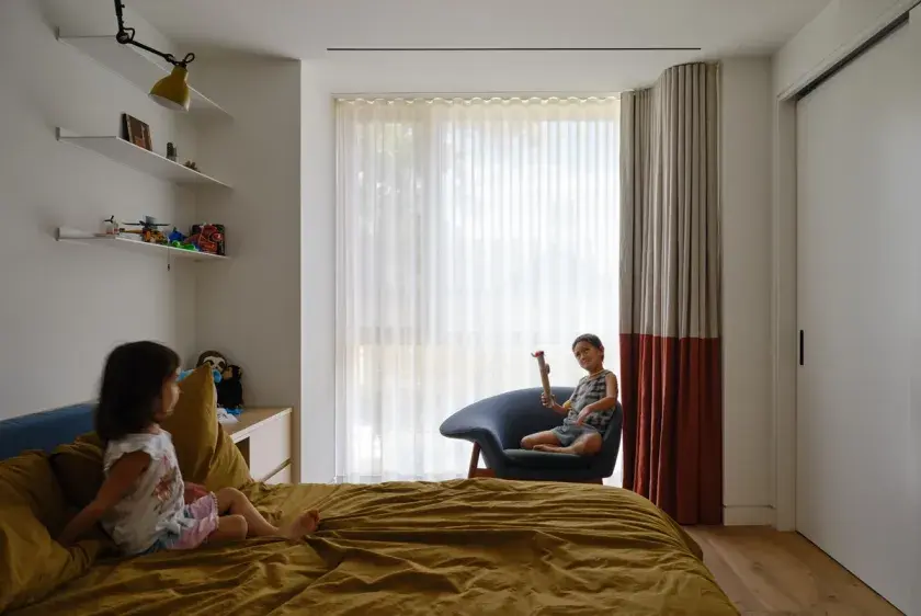 A girl and boy in a child's bedroom. The girl, to the left, sitting on the bed with a mustard duvet, and the boy toward the far right, sits on a blue chair holding a toy.