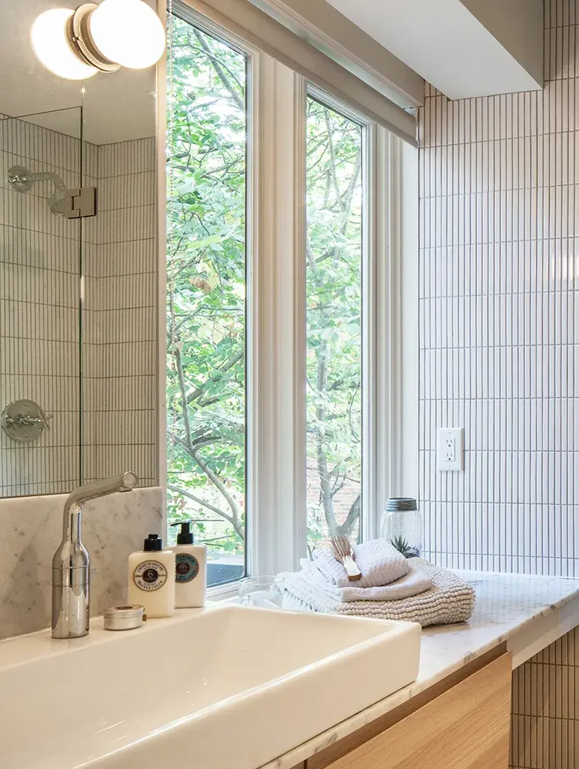 close up of a bathroom with a white vessel sink, white narrow tiles, a marble countertop with folded towels, overlooking the green outdoors