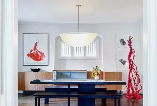 a dining room with a custom walnut banquette, light blue velvet cushions, a black dining table made of glass, a red woven sculpture, and a taco shaped ceiling light