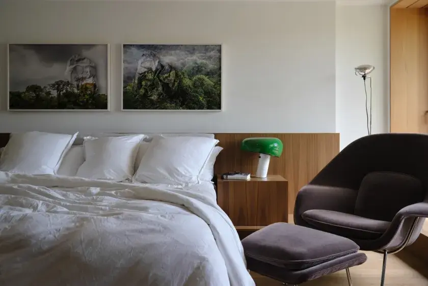 A master bedroom with a white king-size bed, a walnut ‘studiolo’ headboard, a green mushroom side table lamp, and a charcoal velvet lounge chair and ottoman