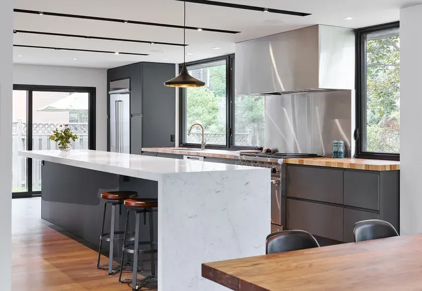 a long single line kitchen with stainless steel appliances, a kitchen island with a light grey countertop, black cabinets, and a wood table in the foreground