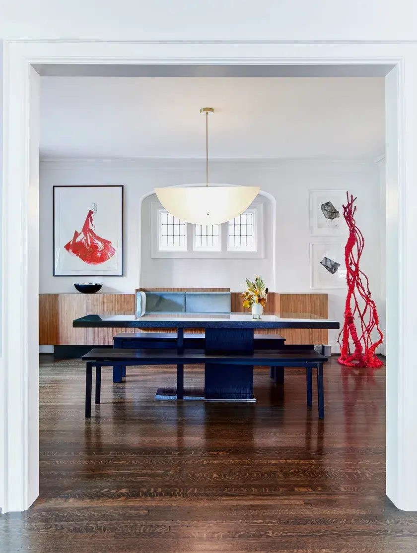 a dining room with a custom walnut banquette, light blue velvet cushions, a black dining table made of glass, a red woven sculpture, and a taco shaped ceiling light