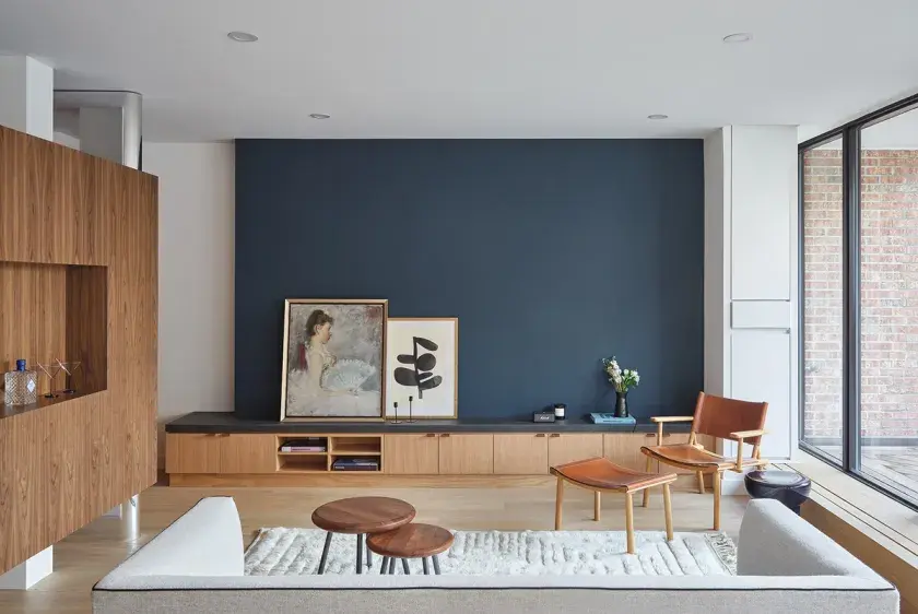 A low custom oak credenza against a navy blue accent wall, and a custom walnut floating wall to the left. The room is furnished with a grey couch facing the blue accent wall, and a caramel leather seat faces the viewer