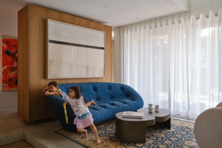 Two children in a living room sitting on a royal blue, cloud-like sofa with a blackened metal coffee table in the foreground, custom walnut floating cabinet in the background and floor-to-ceiling sheer curtains to the right.