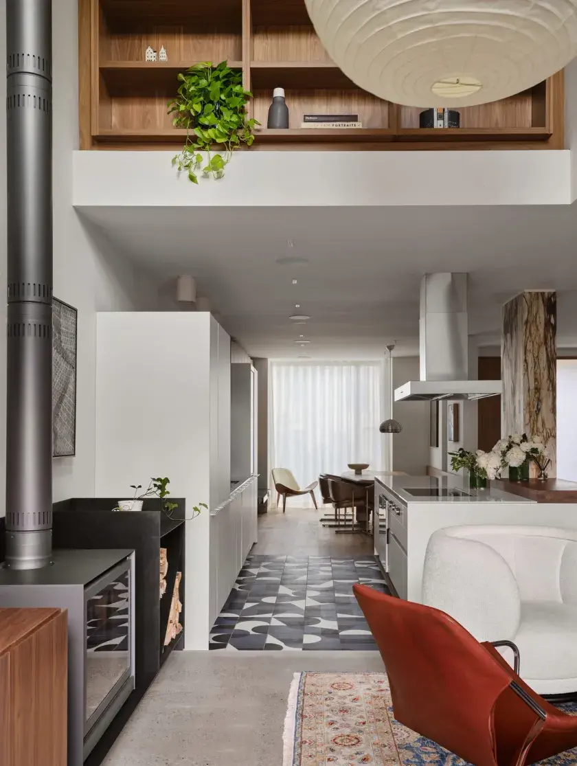 A portrait of a long floorplan with a double-height ceiling that connects living, kitchen, and dining rooms. Materials include marble slabs clad steps, counters, benches and posts, warming concrete flooring, and custom walnut millwork.