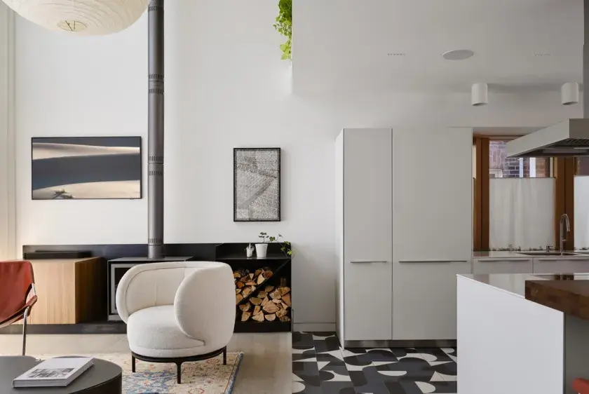 The living and kitchen areas meeting. To the left, a white accent chair and modern mid-century fireplace, sitting on warming concrete flooring. To the right, a white kitchen pantry and marble island, resting above graphic floor tiling.