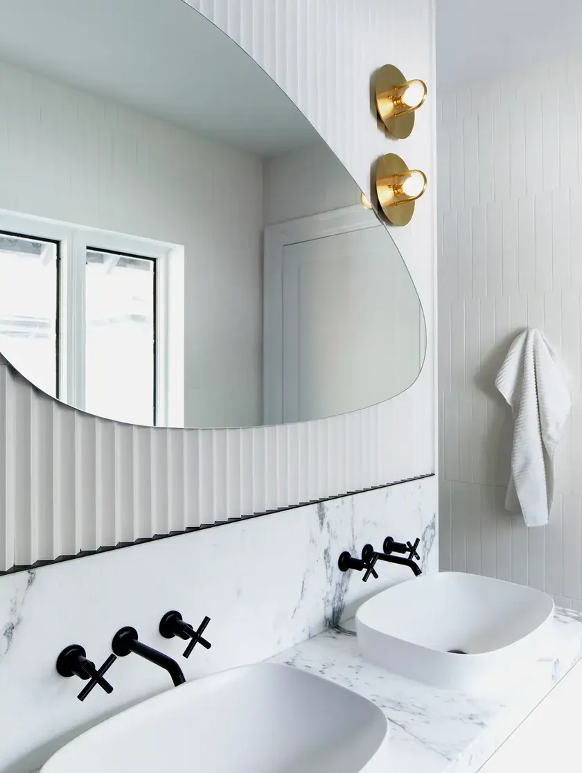 a his-and hers bathroom with two white vessel sinks, on top of a light grey quartz, and off-white bevelled backsplash, black fixtures and gold sconces