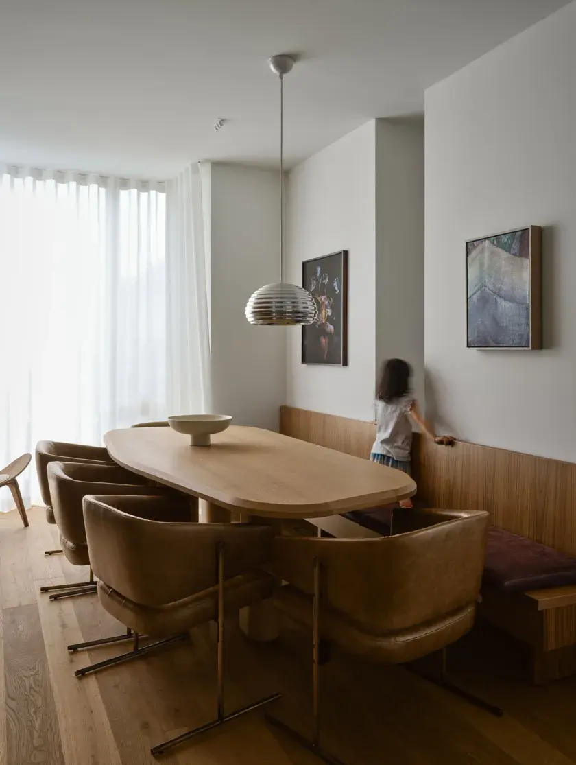 A dining room with 4 leather chair placed around a wood dining table and chrome ceiling light fixture. To the right, a custom walnut banquet acts as an accent wall. A little girl peers through a divided wall behind it.