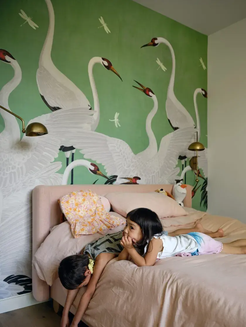Two children laying on a bed with a light pink duvet in front of a Gucci accent wallpaper featuring a light green background and white herons
