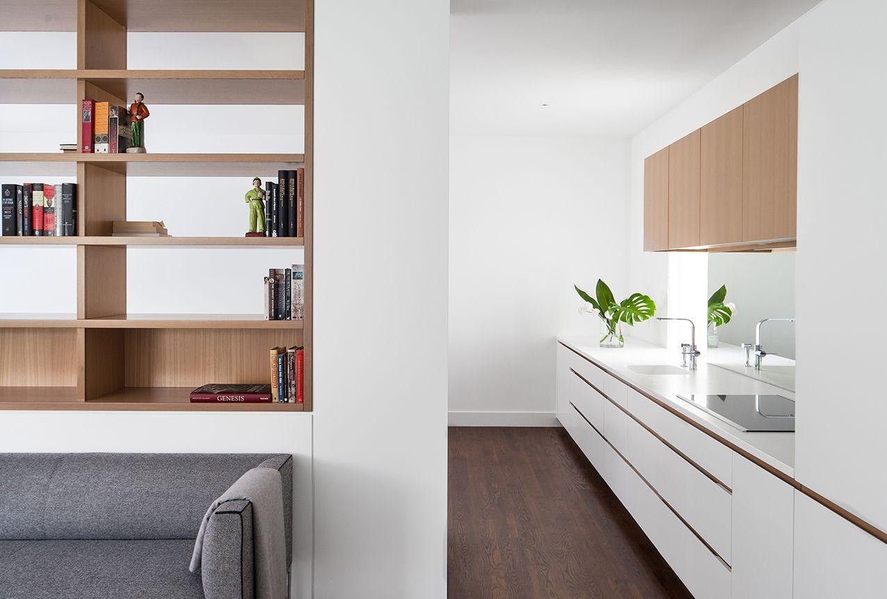close up of a custom millwork bookshelf above a grey couch, next to a white single line kitchen with a mirrored backsplash