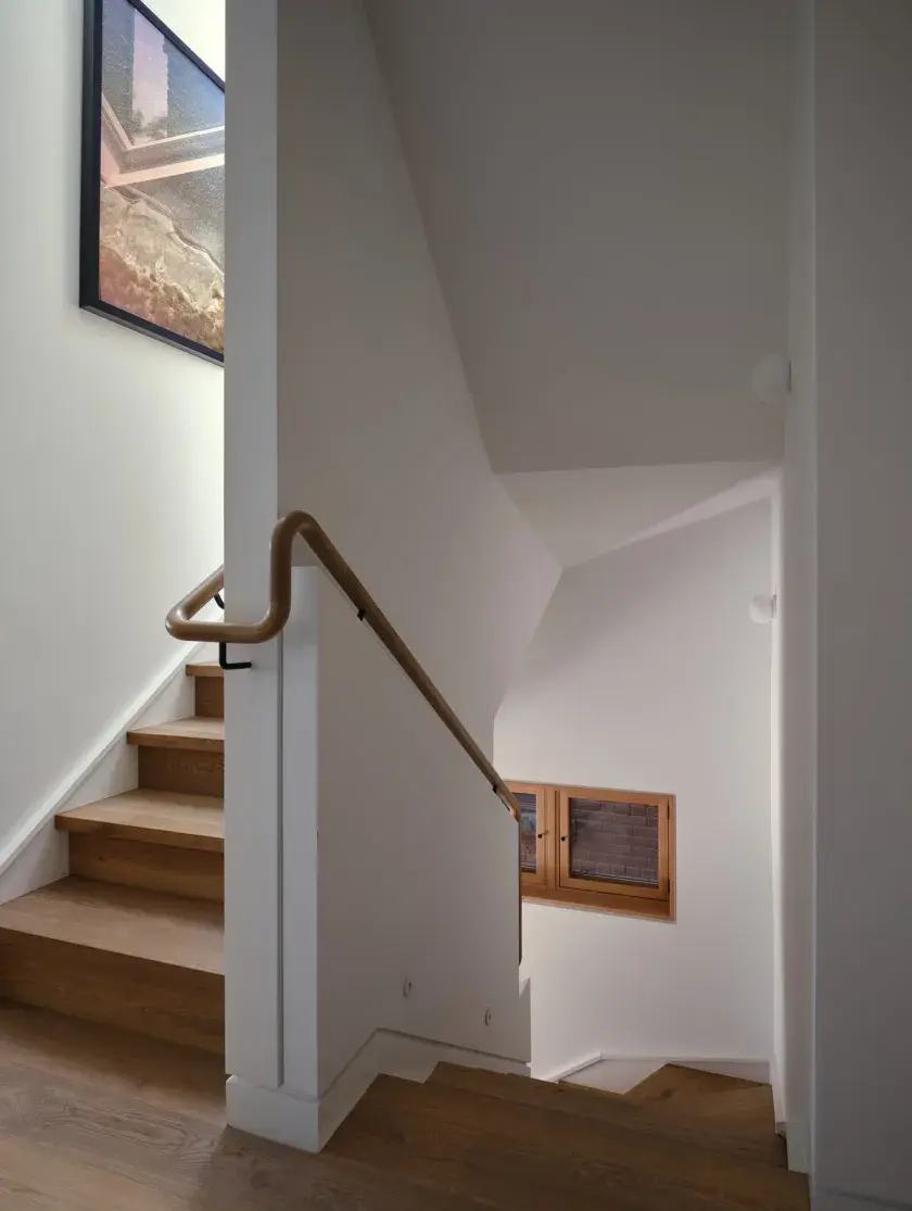 A landing of the upper level staircase showing the ascending flight to the left, and descending flight to the right. A smooth, sculpted continuous white oak wood handrail acts as guide for users.