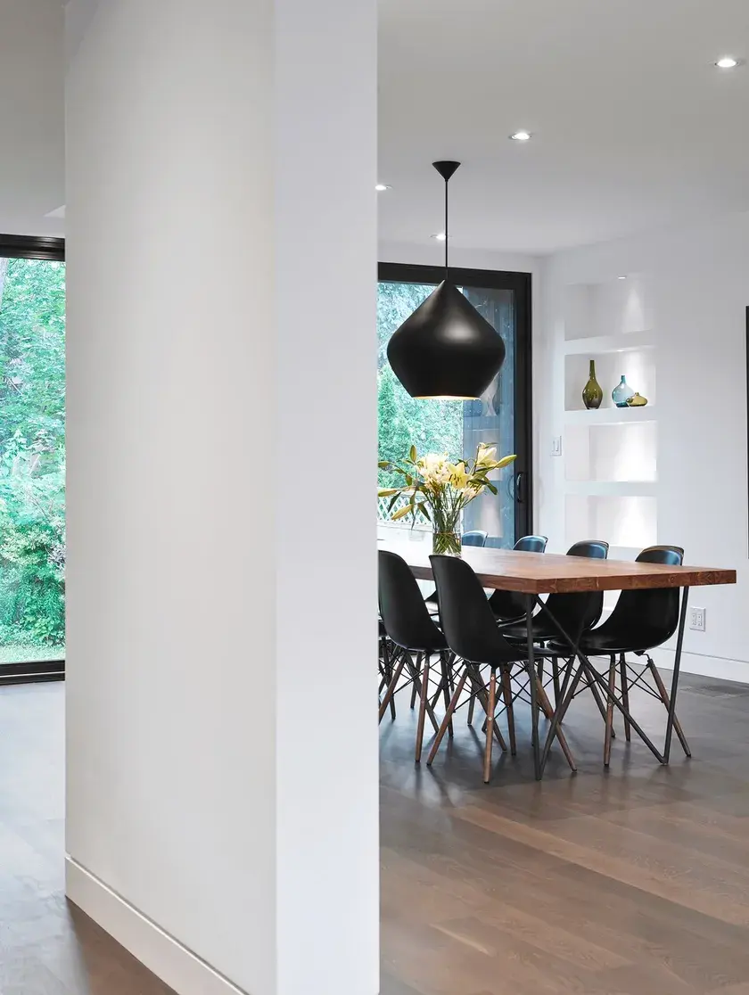 a white dividing wall in the foreground, and a dining room in the background with dark smoked oak floors, a wood table and six black eames chairs by the large sliding doors overlooking the backyard
