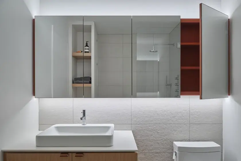 A straight on view of a wood bathroom vanity, with warm grey tiled backsplash, wall-to-wall mirror cabinets and one cabinet door opened to expose red interior finishes