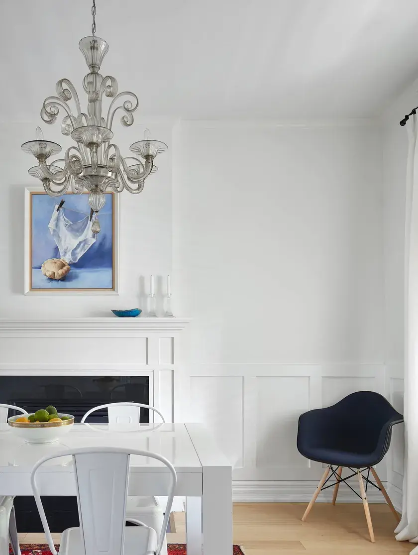 close up of an all white dining room with a silver chandelier, white fireplace, and navy blue eames chair
