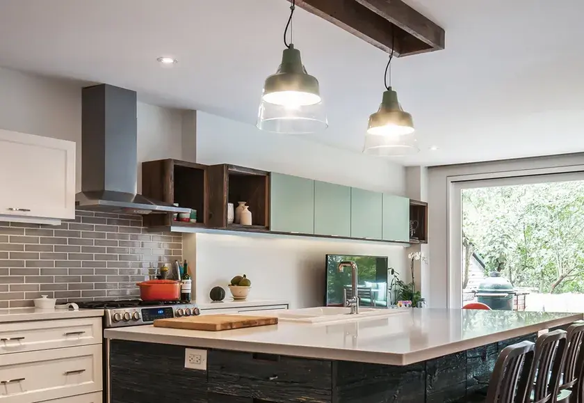 a single-line kitchen with grey subway tiles, a stainless steel vent hood, a white island countertop with dark raw wood finishing, and industrial lights