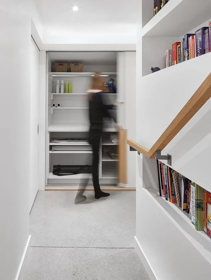 The entryway to a lower level hallway with a tall woman opening a custom millwork laundry cabinet, polished concrete floors, and a built in bookshelf at the end of the staircase